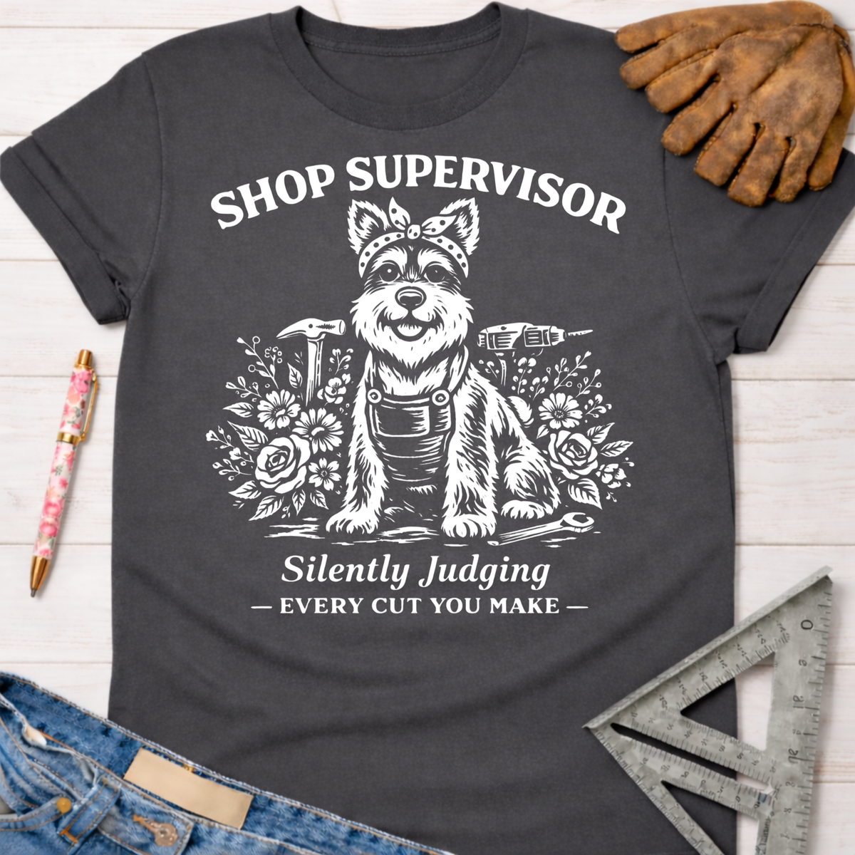 Women's funny woodworking graphic tee shirt for female woodworkers, makers, and DIY builders — Sawdust & Ponytails. Dark gray t-shirt with a white graphic of a dog wearing a bandana and overalls, surrounded by tools and flowers, with text "Shop Supervisor Silently Judging Every Cut You Make.