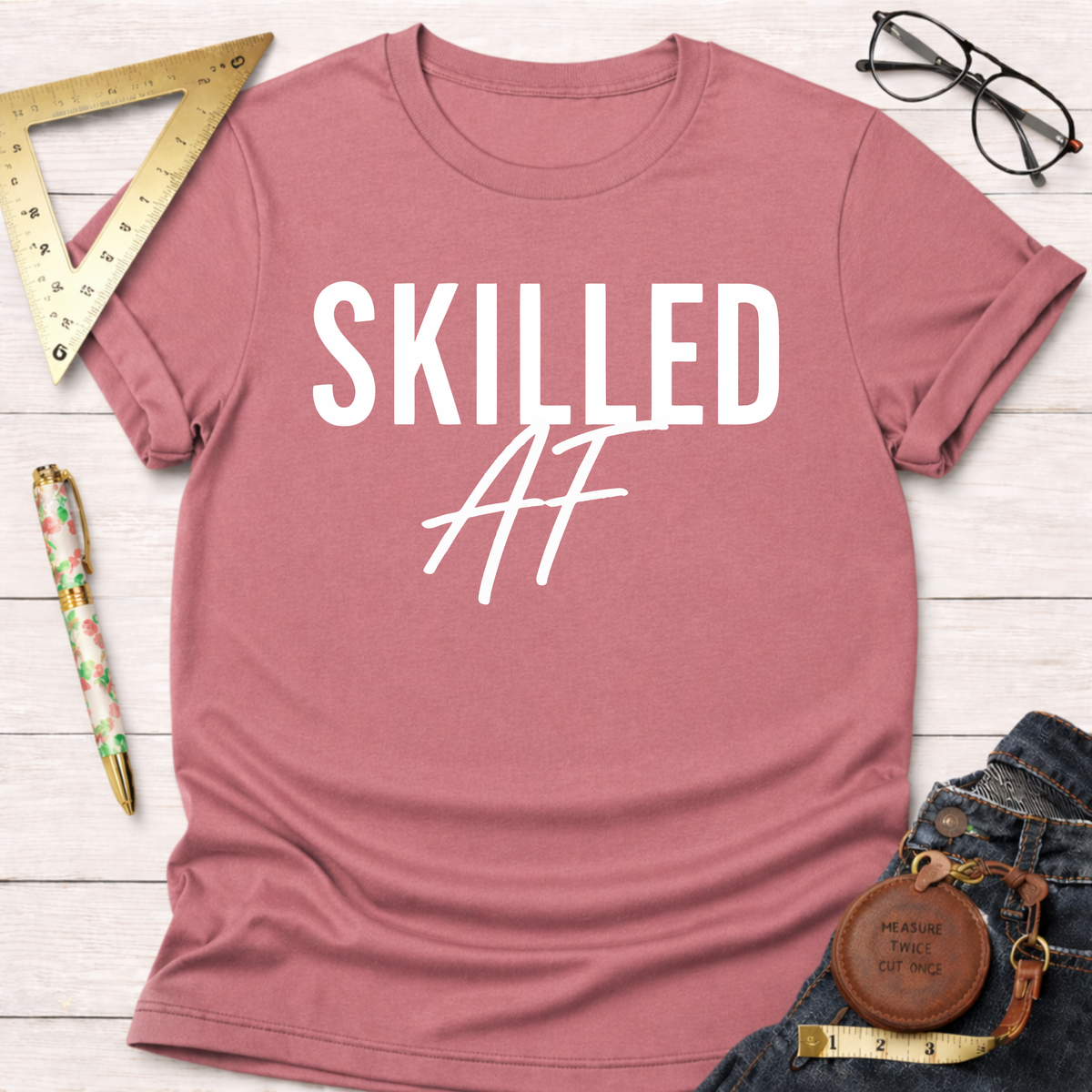 Skilled AF T-shirt in dusty rose pink cotton with white text, styled with jeans and workshop tools.
