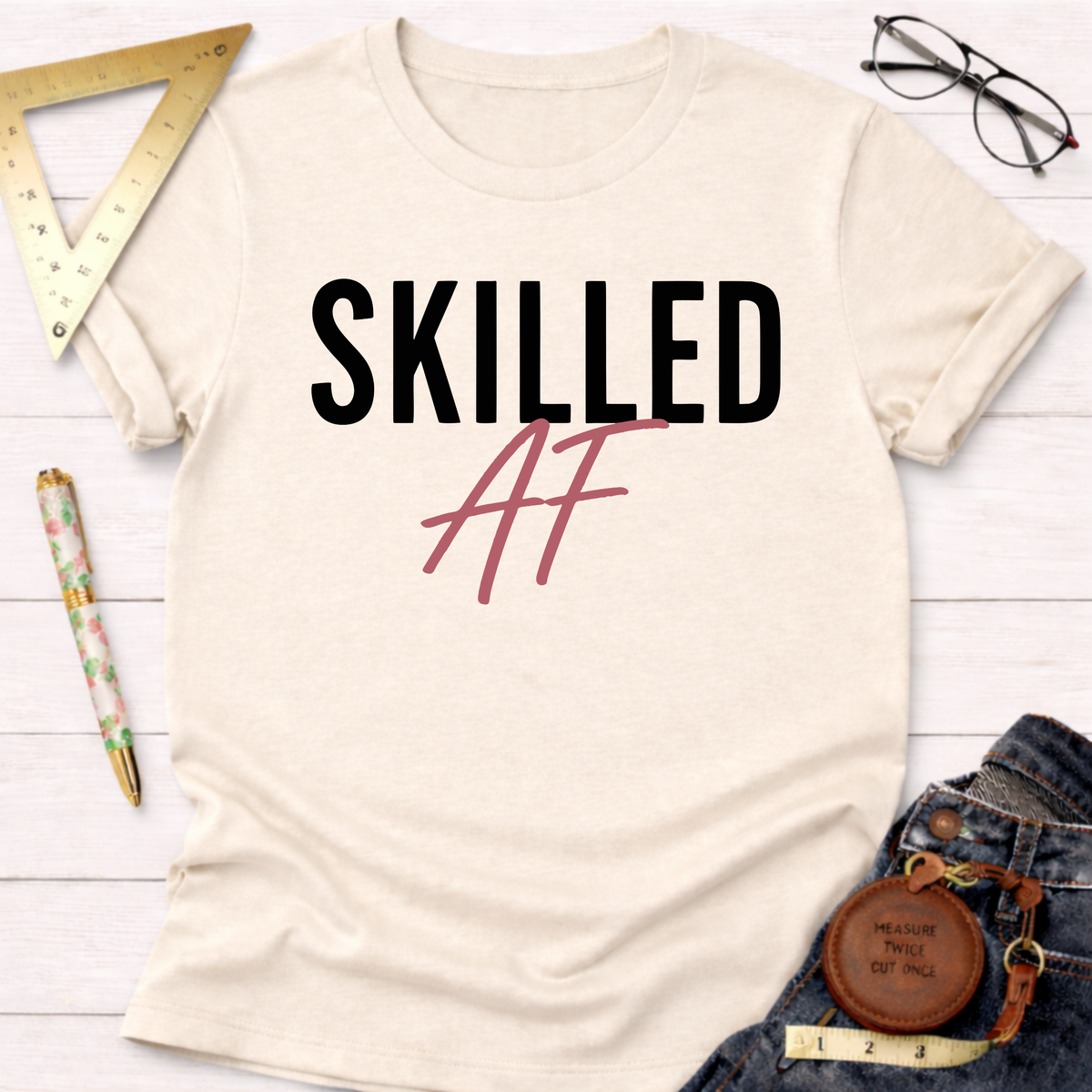 Cream cotton "SKILLED AF" t-shirt with black and pink text, styled with jeans and tools.