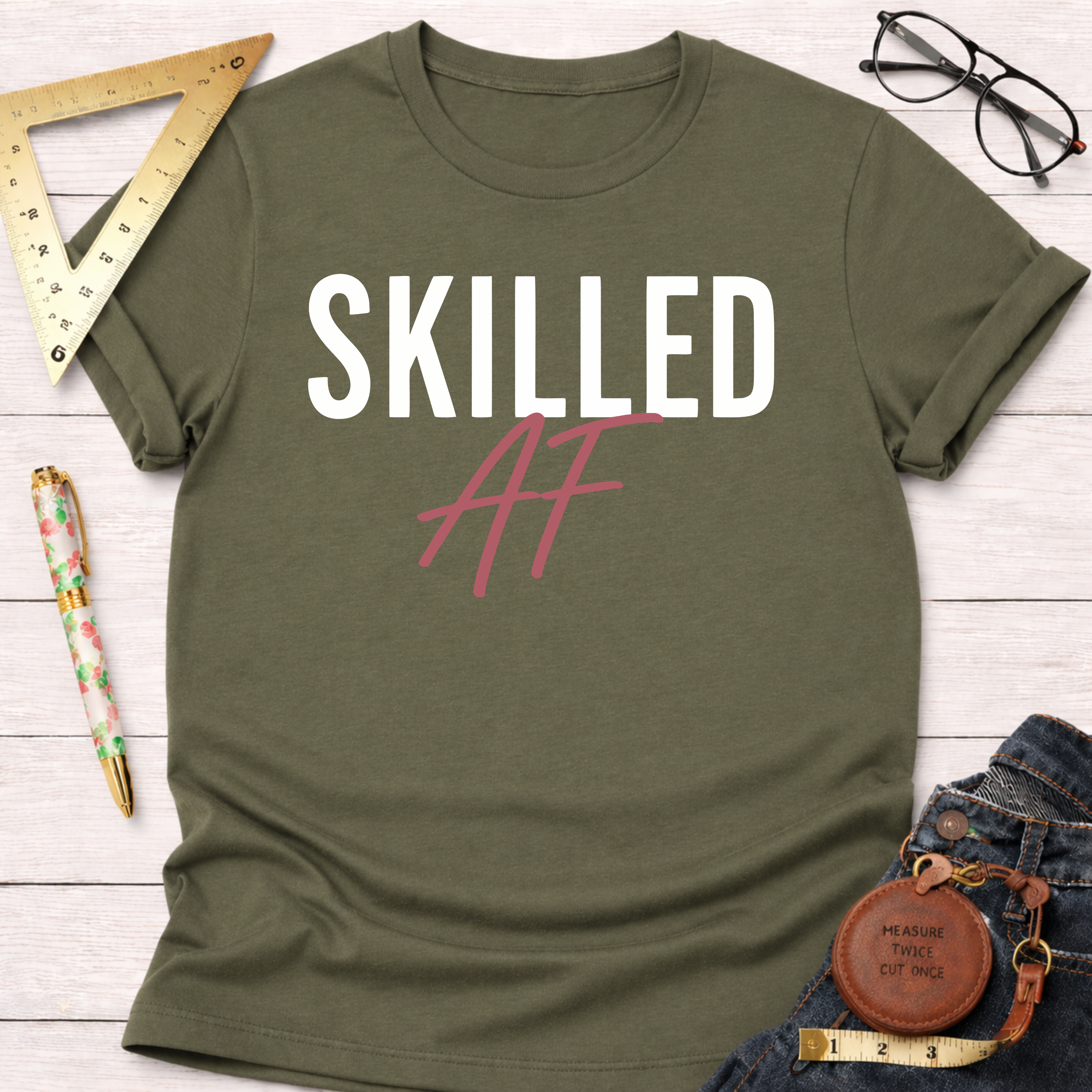 Olive green cotton t-shirt with "SKILLED AF" in white and pink, folded on a white wooden surface.