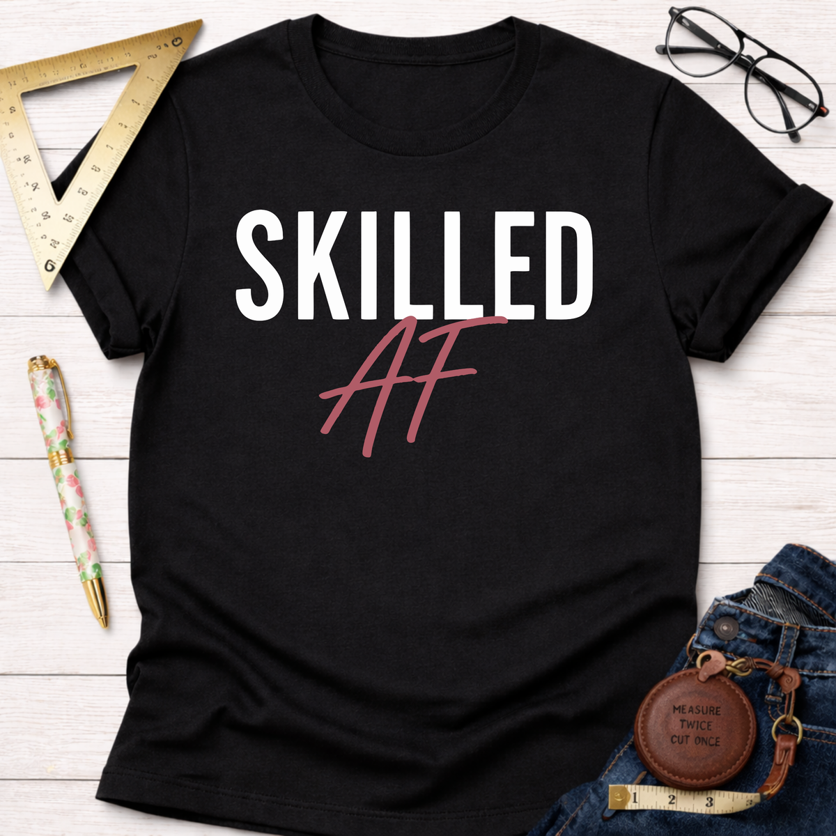 Black cotton t-shirt with "Skilled AF" in white and pink, styled with jeans, ruler, and glasses.