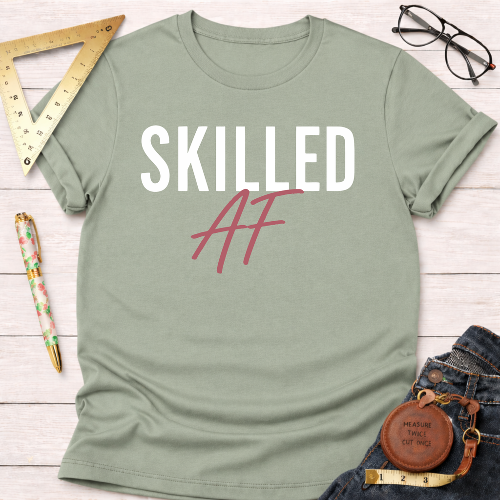 Sage green "SKILLED AF" t-shirt, folded on a white wooden surface with tools and jeans.