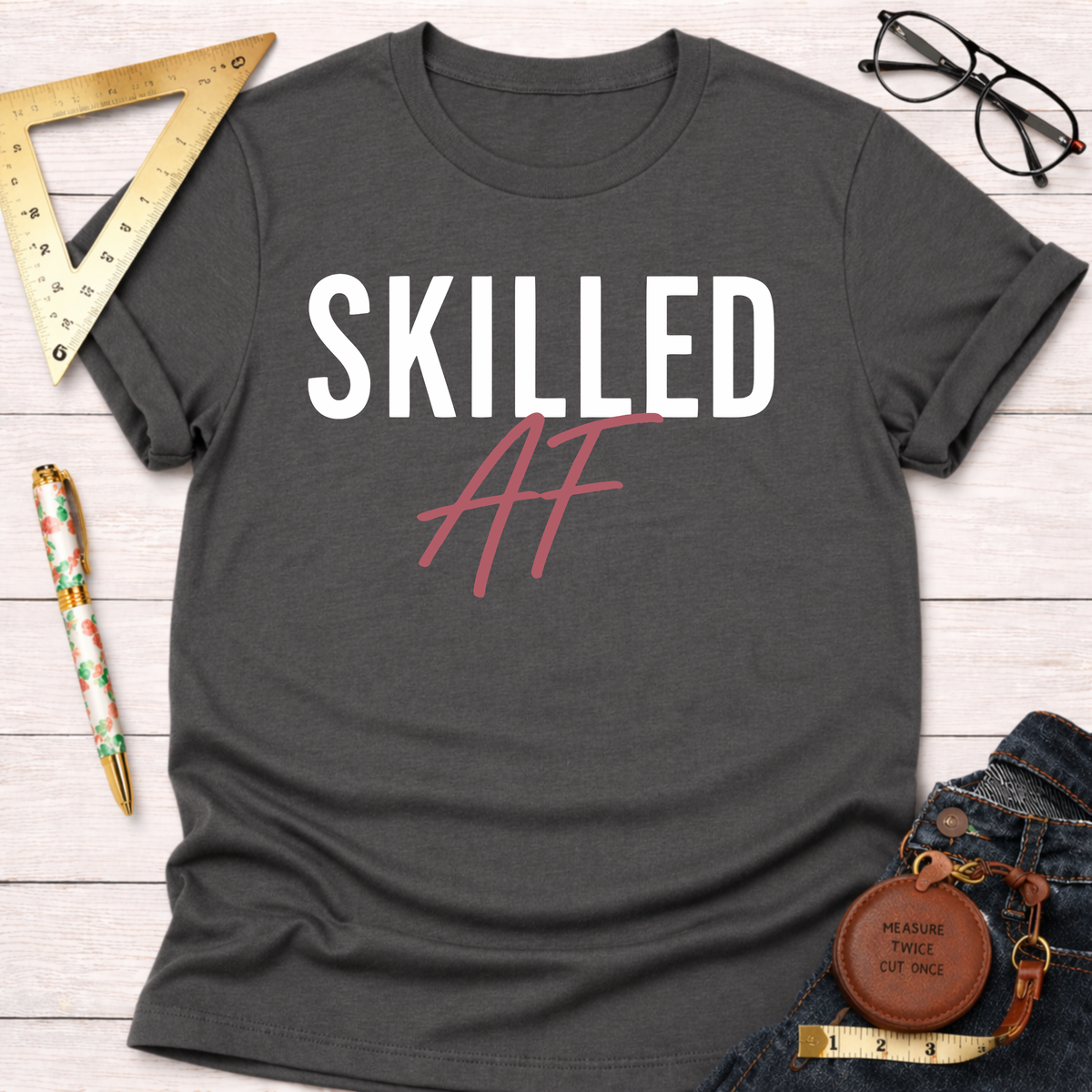 Charcoal gray cotton t-shirt with "SKILLED AF" in white and pink, styled with jeans and tools.