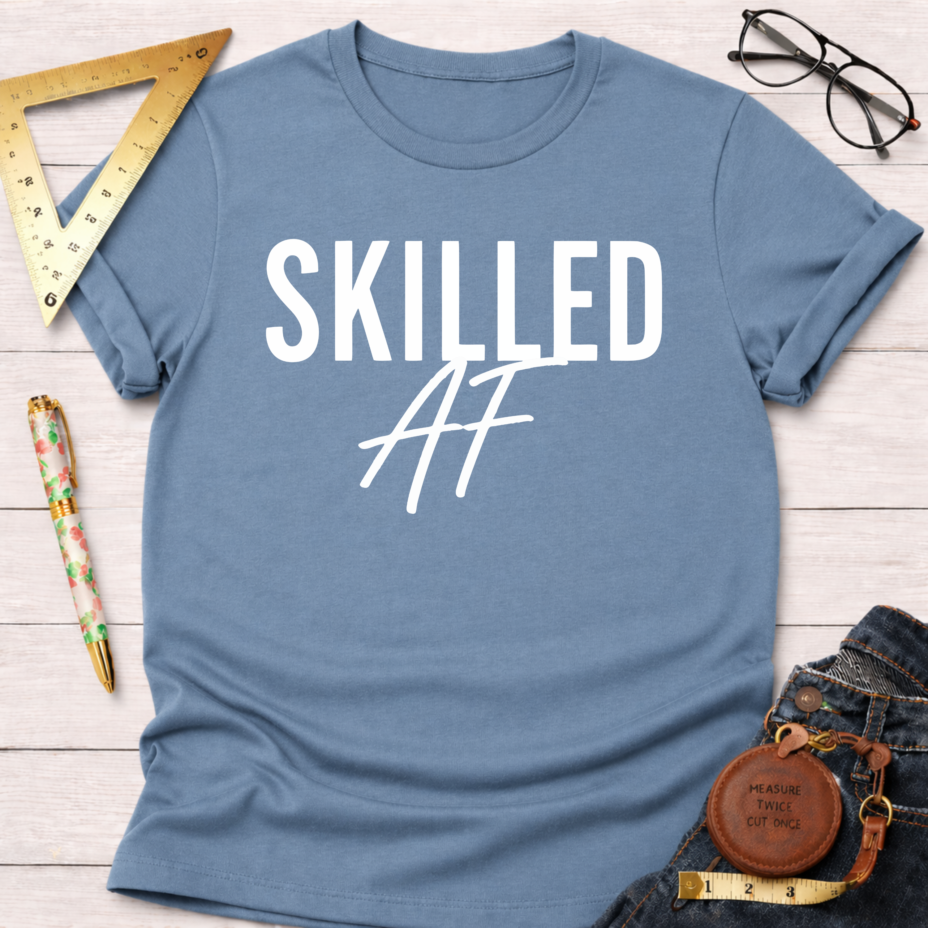 Blue cotton "Skilled AF" t-shirt, flat lay with jeans, ruler, pen, and glasses.