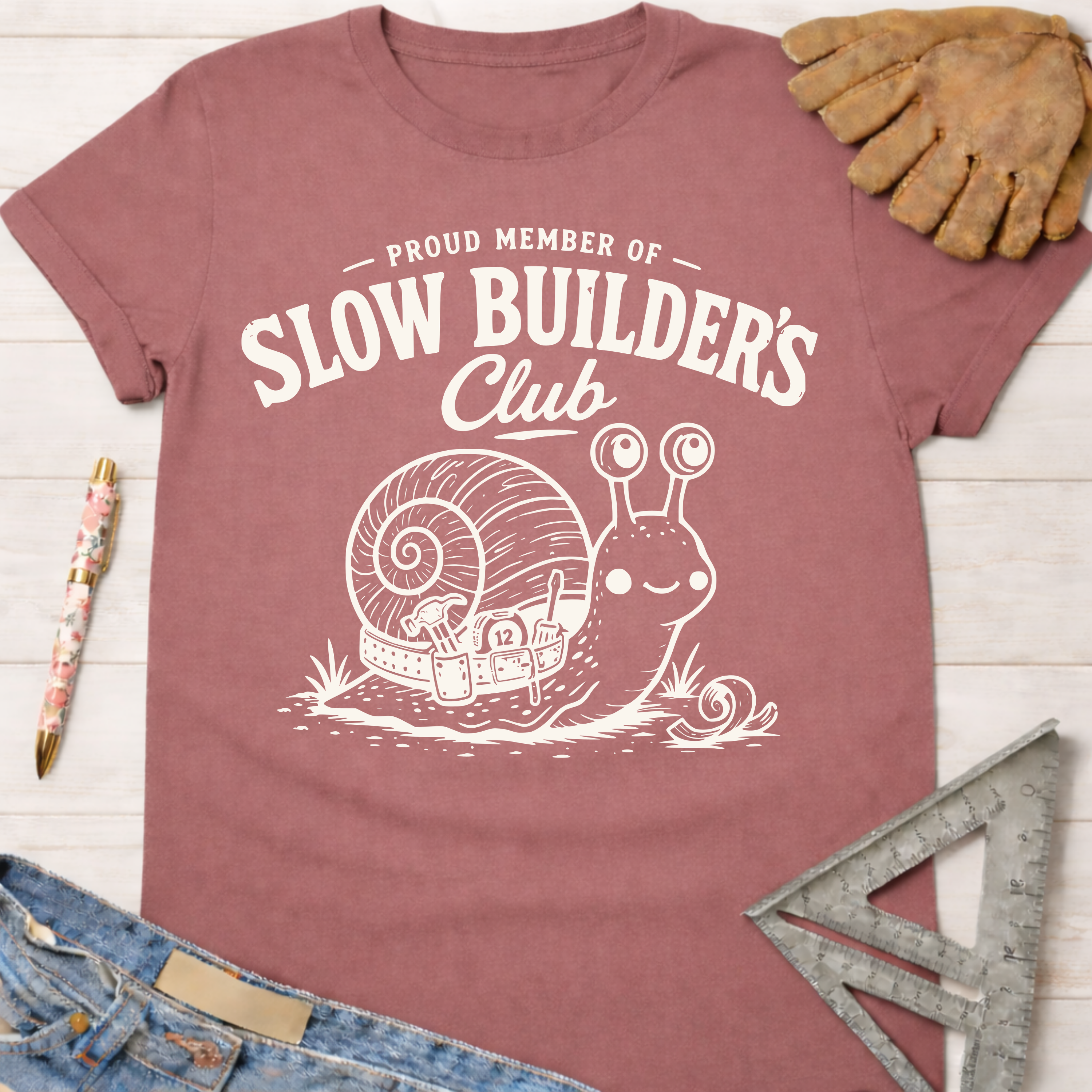 Women's funny woodworking graphic tee shirt for female woodworkers, makers, and DIY builders — Sawdust & Ponytails. Mauve t-shirt with "Slow Builder's Club" text and a cute snail pulling a boat.