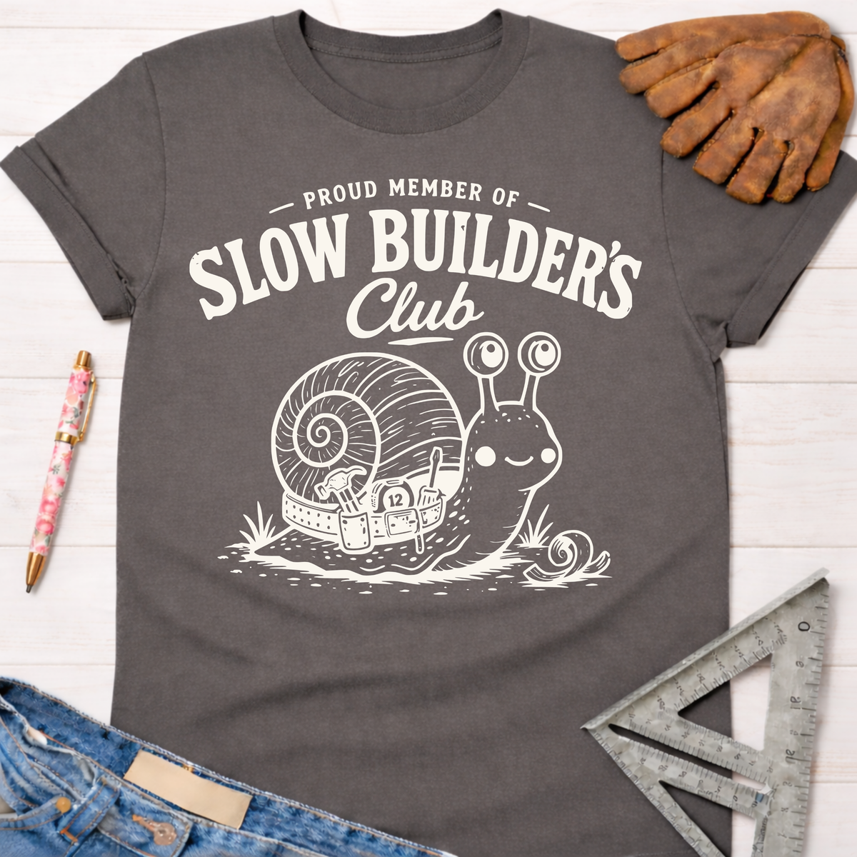 Women's funny woodworking graphic tee shirt for female woodworkers, makers, and DIY builders — Sawdust & Ponytails. Dark gray t-shirt with "Slow Builder's Club" text and a happy snail carrying tools.