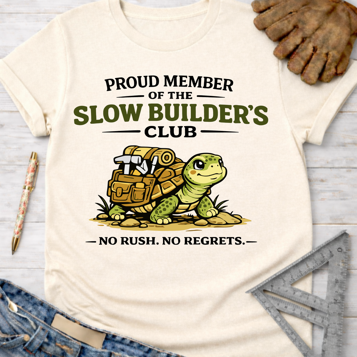 Women's funny woodworking graphic tee shirt for female woodworkers, makers, and DIY builders — Sawdust & Ponytails. Cream t-shirt with "Proud Member of the Slow Builder's Club" text and a turtle carrying tools.