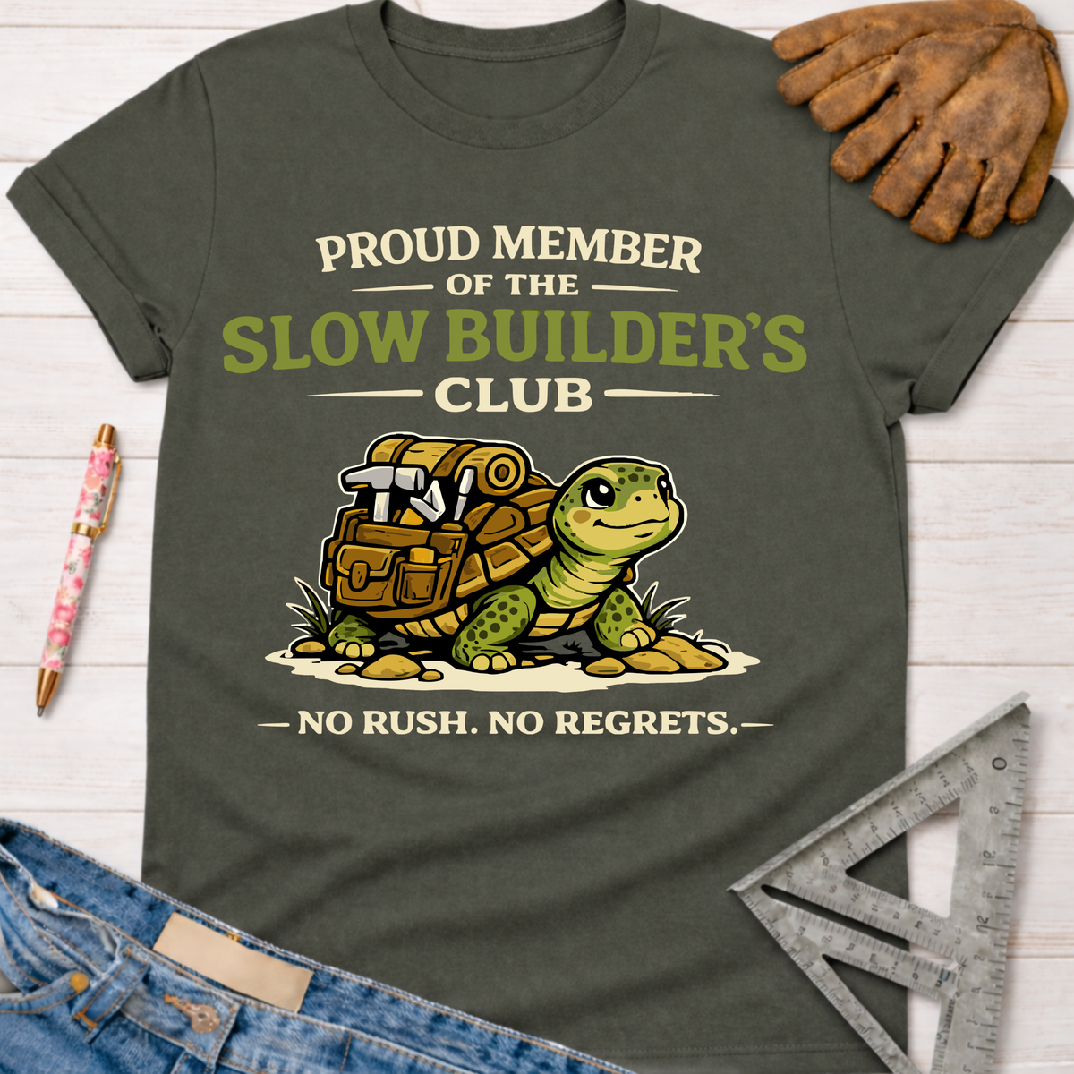 Women's funny woodworking graphic tee shirt for female woodworkers, makers, and DIY builders — Sawdust & Ponytails. Dark olive green cotton tee with a cartoon turtle carrying tools and text "Proud Member of the Slow Builder's Club - No Rush. No Regrets.