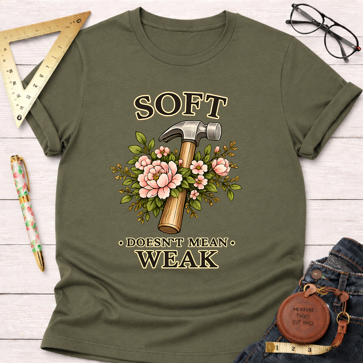 Olive green t-shirt with "SOFT DOESN'T MEAN WEAK" text, featuring a hammer surrounded by pink flowers.