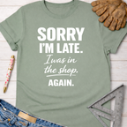 Women's funny woodworking graphic tee shirt for female woodworkers, makers, and DIY builders — Sawdust & Ponytails. Sage green cotton t-shirt with "Sorry I'm Late. I was in the shop. Again." in white text.