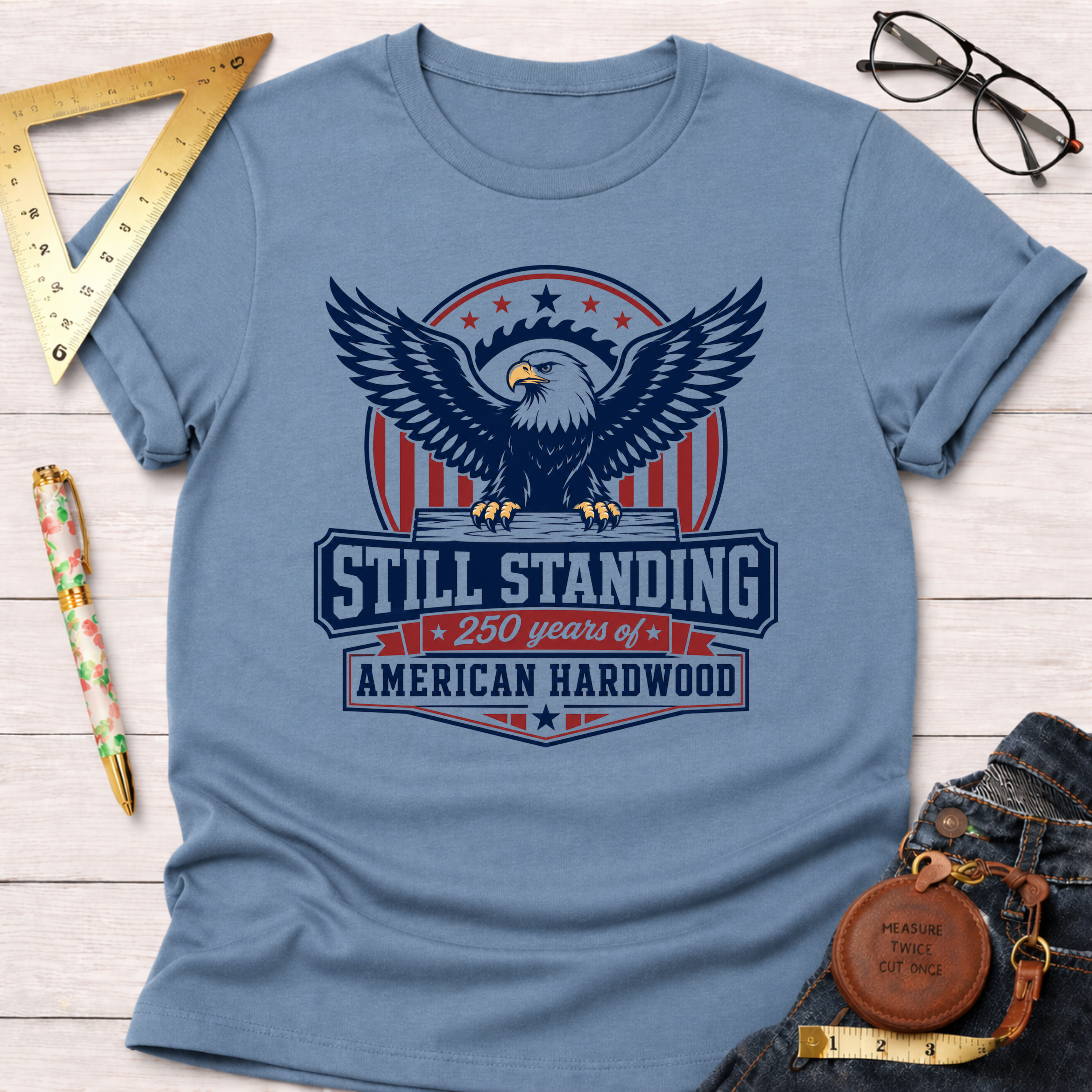 Light blue cotton t-shirt with a "Still Standing American Hardwood" eagle graphic, styled with jeans and tools.