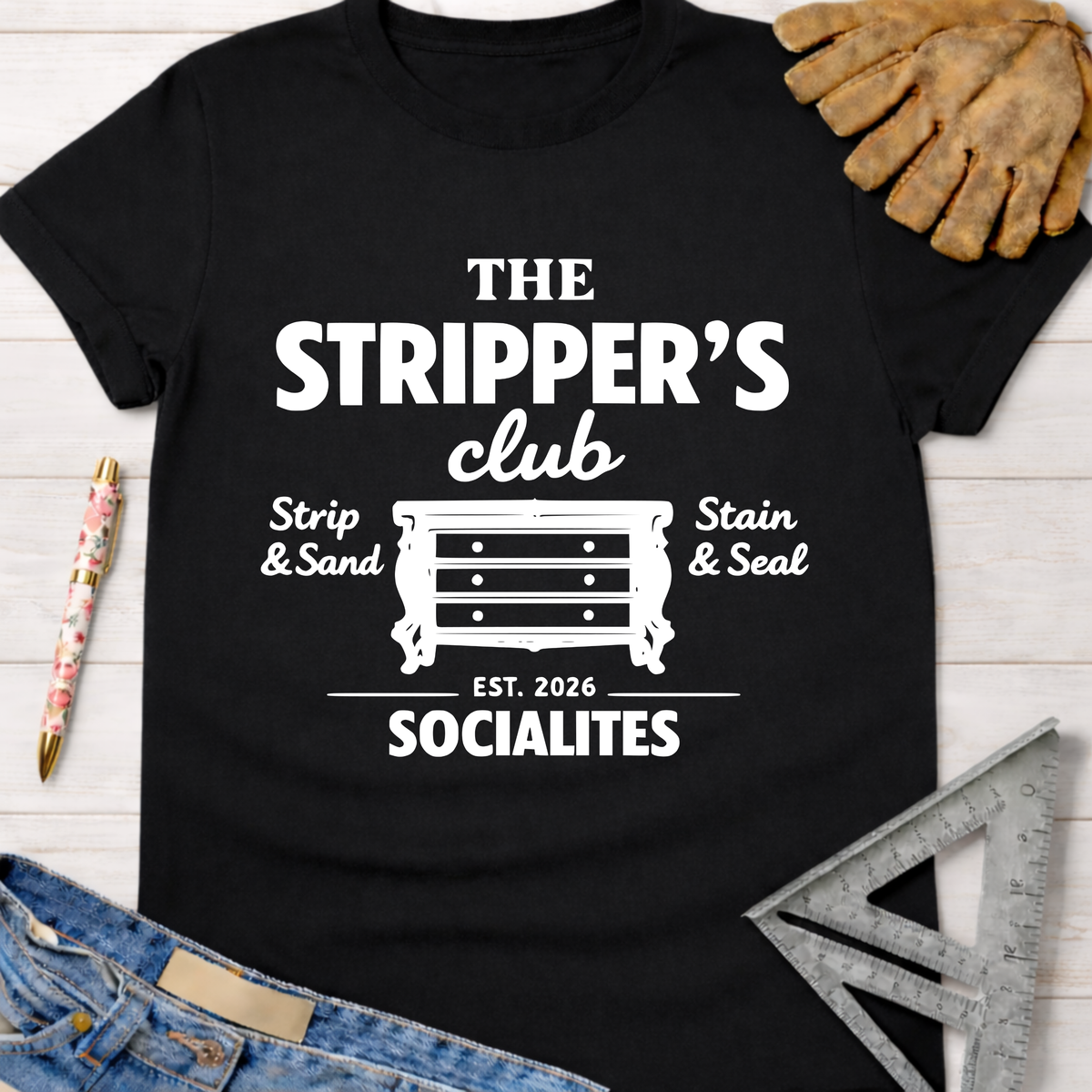 Black cotton tee with "The Stripper's Club Socialites" graphic, dresser icon, and "Strip & Sand, Stain & Seal." Women's funny woodworking graphic tee shirt for female woodworkers, makers, and DIY builders — Sawdust & Ponytails.