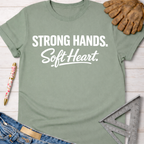Women's funny woodworking graphic tee shirt for female woodworkers, makers, and DIY builders — Sawdust & Ponytails. Sage green cotton tee with "Strong Hands. Soft Heart." in white text.