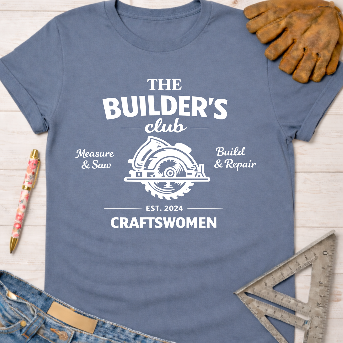 Women's funny woodworking graphic tee shirt for female woodworkers, makers, and DIY builders — Sawdust & Ponytails. Blue cotton tee with "The Builder's Club" graphic, circular saw, and "Craftswomen Est. 2024".