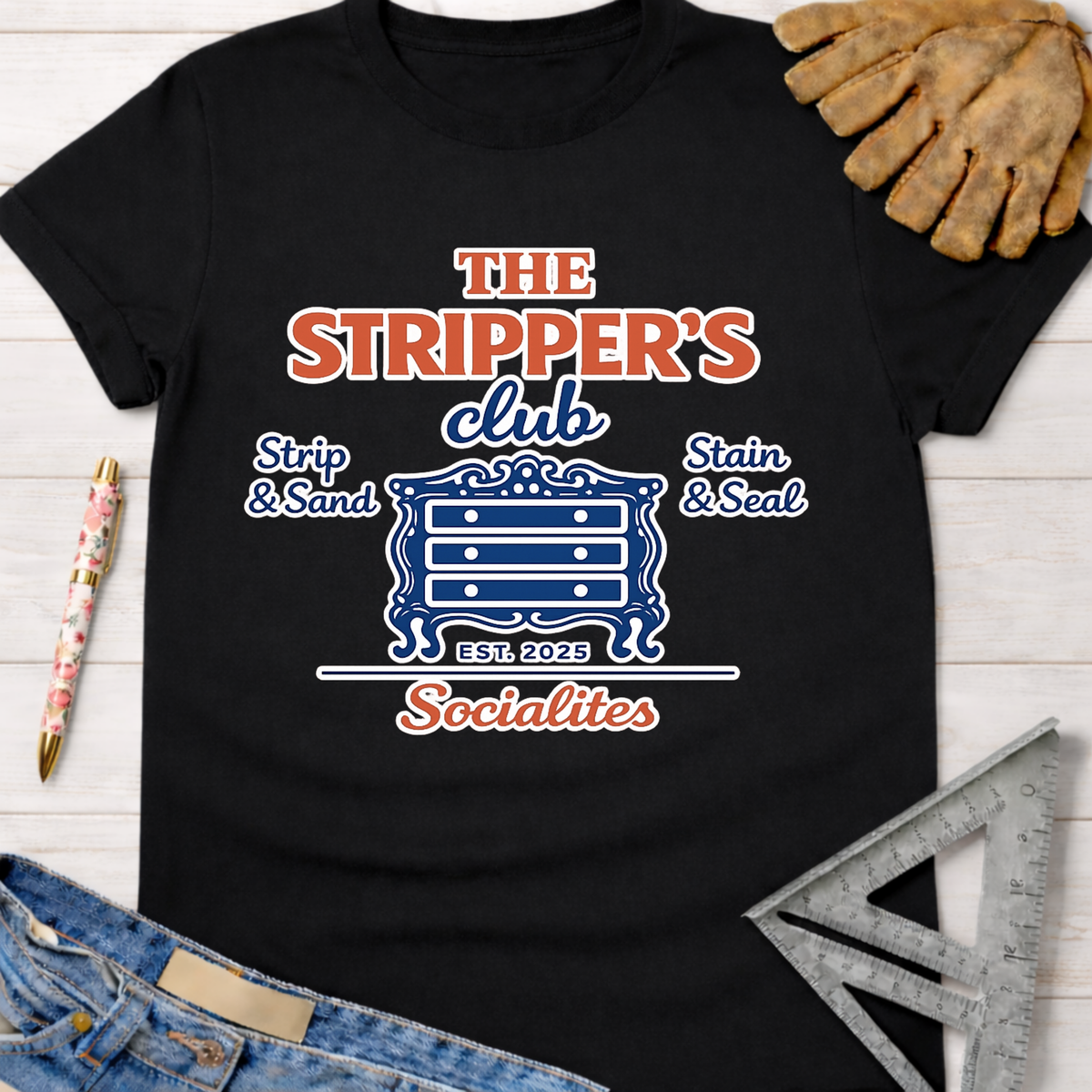 Black cotton tee with "The Stripper's Club Socialites" graphic, dresser icon, and "Strip & Sand, Stain & Seal" text. Women's funny woodworking graphic tee shirt for female woodworkers, makers, and DIY builders — Sawdust & Ponytails