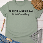 Women's funny woodworking graphic tee shirt for female woodworkers, makers, and DIY builders — Sawdust & Ponytails. Sage green cotton t-shirt with "TODAY IS A GOOD DAY to build something" text, shown with jeans, work gloves, and a ruler.