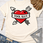 Women's funny woodworking graphic tee shirt for female woodworkers, makers, and DIY builders — Sawdust & Ponytails. Cream t-shirt with a red heart, arrows, and "TOOL SLUT" banner design.