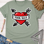 Women's funny woodworking graphic tee shirt for female woodworkers, makers, and DIY builders — Sawdust & Ponytails. Sage green t-shirt with a red heart, black arrows, and "TOOL SLUT" banner.