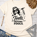 Women's funny woodworking graphic tee shirt for female woodworkers, makers, and DIY builders — Sawdust & Ponytails. Cream t-shirt with "Tools Before Fools" graphic featuring a retro woman holding a hammer.