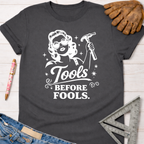 Women's funny woodworking graphic tee shirt for female woodworkers, makers, and DIY builders — Sawdust & Ponytails. Dark gray cotton tee with "Tools Before Fools" text and a retro woman holding a hammer.