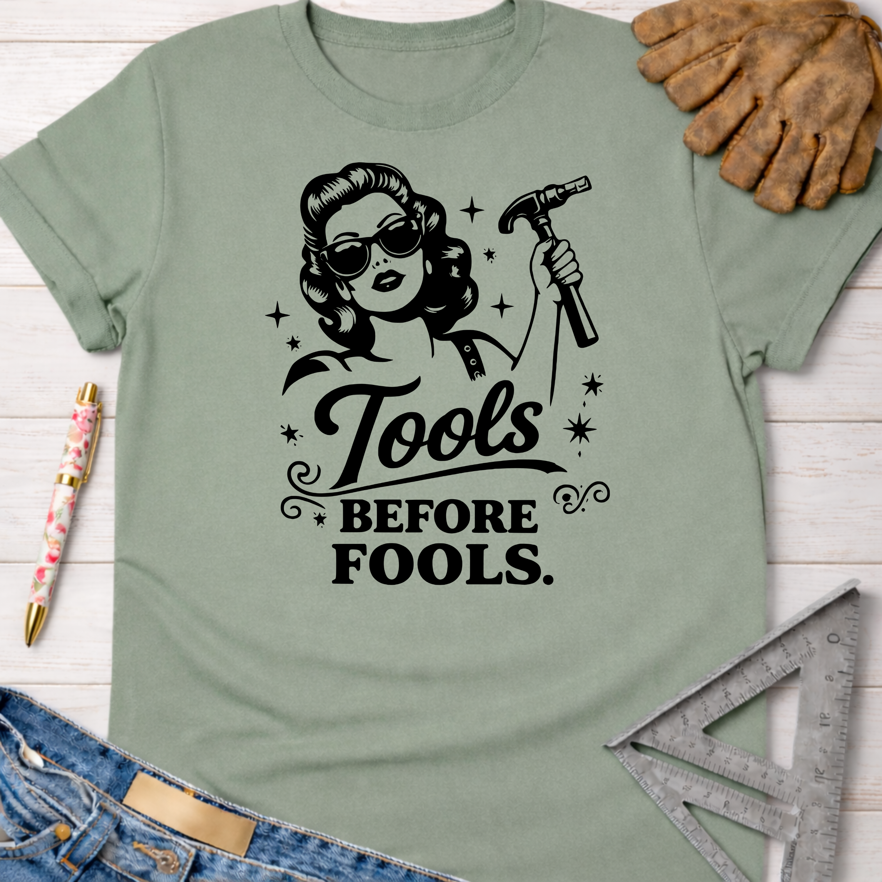 Women's funny woodworking graphic tee shirt for female woodworkers, makers, and DIY builders — Sawdust & Ponytails. Sage green t-shirt with "Tools Before Fools" graphic featuring a retro woman holding a hammer.