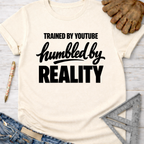Women's funny woodworking graphic tee shirt for female woodworkers, makers, and DIY builders — Sawdust & Ponytails. Cream t-shirt with "Trained By YouTube Humbled By Reality" in black text.