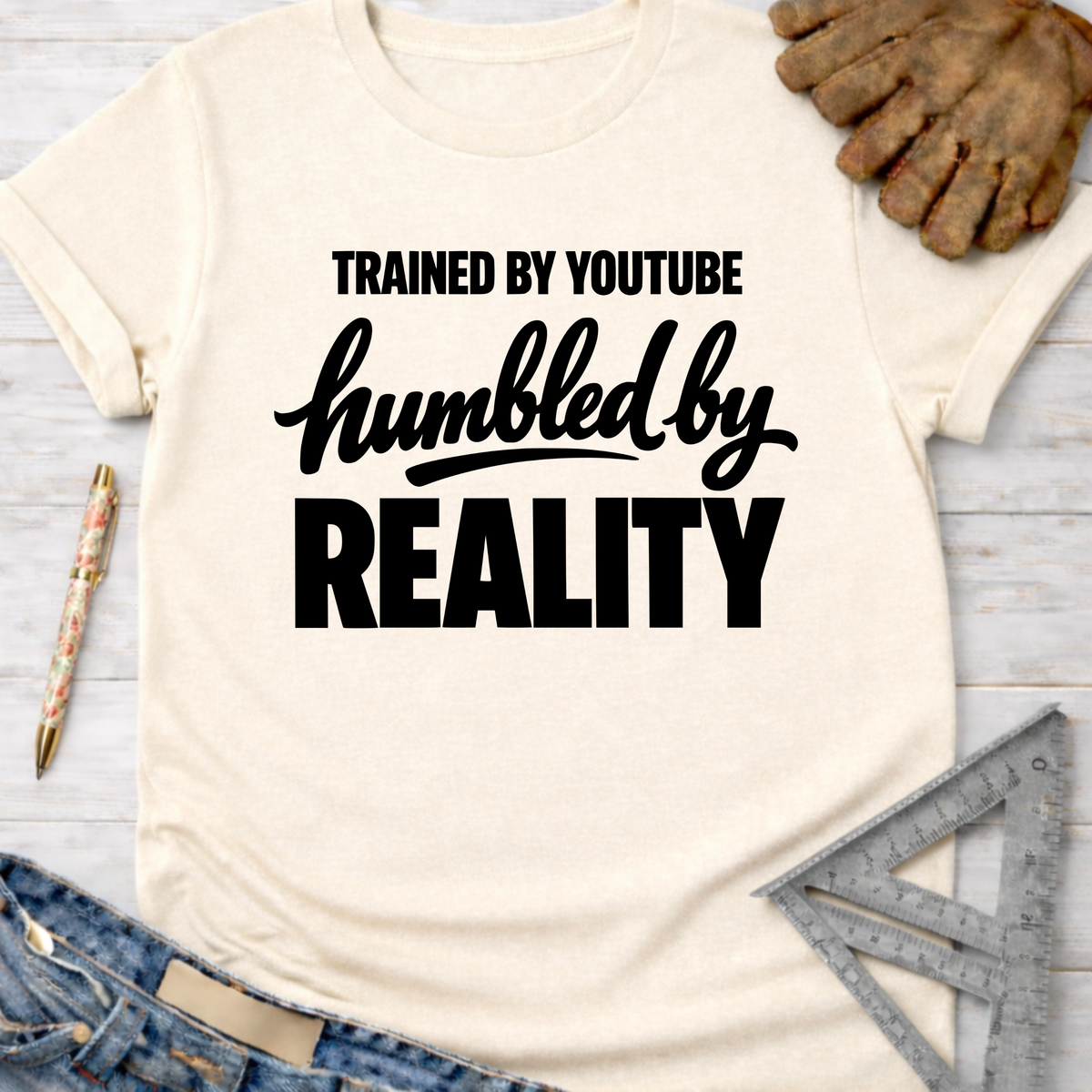 Women's funny woodworking graphic tee shirt for female woodworkers, makers, and DIY builders — Sawdust & Ponytails. Cream t-shirt with "Trained By YouTube Humbled By Reality" in black text.