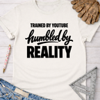 Women's funny woodworking graphic tee shirt for female woodworkers, makers, and DIY builders — Sawdust & Ponytails. White cotton tee with "Trained By YouTube Humbled By REALITY" in black text.