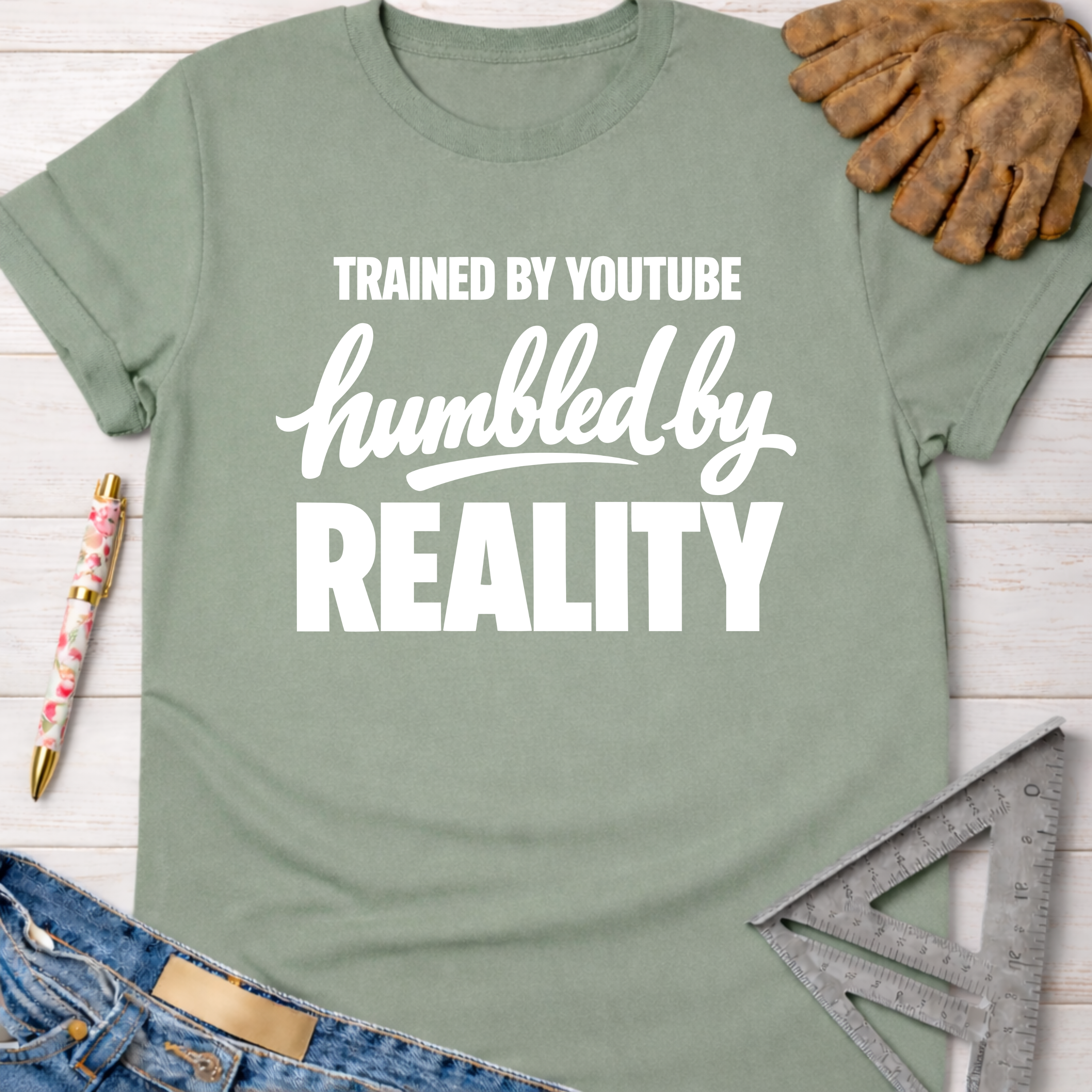 Women's funny woodworking graphic tee shirt for female woodworkers, makers, and DIY builders — Sawdust & Ponytails. Sage green cotton tee with "Trained By YouTube Humbled By Reality" in white text.