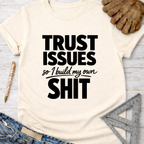 Women's funny woodworking graphic tee shirt for female woodworkers, makers, and DIY builders — Sawdust & Ponytails. Cream t-shirt with "TRUST ISSUES so I build my own SHIT" in black text.