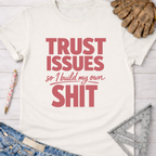 Women's funny woodworking graphic tee shirt for female woodworkers, makers, and DIY builders — Sawdust & Ponytails. Cream t-shirt with "TRUST ISSUES so I build my own SHIT" in red text.