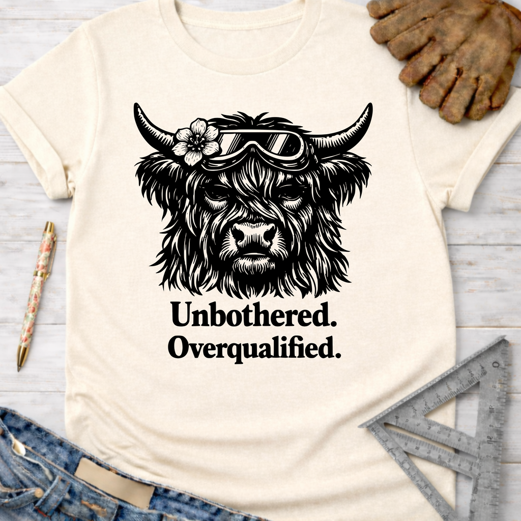 Women's funny woodworking graphic tee shirt for female woodworkers, makers, and DIY builders — Sawdust & Ponytails. Cream t-shirt with a black graphic of a Highland cow wearing goggles and a flower, text "Unbothered. Overqualified.