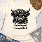 Women's funny woodworking graphic tee shirt for female woodworkers, makers, and DIY builders — Sawdust & Ponytails. Cream t-shirt with a black graphic of a Highland cow wearing goggles and a flower, text "Unbothered. Overqualified.