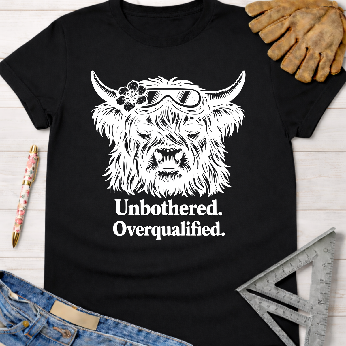 Women's funny woodworking graphic tee shirt for female woodworkers, makers, and DIY builders — Sawdust & Ponytails. Black cotton tee with white graphic of a Highland cow wearing goggles and a flower, text "Unbothered. Overqualified.