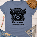 Women's funny woodworking graphic tee shirt for female woodworkers, makers, and DIY builders — Sawdust & Ponytails. Blue tee with "Unbothered. Overqualified." text and a Highland cow wearing goggles and a flower.