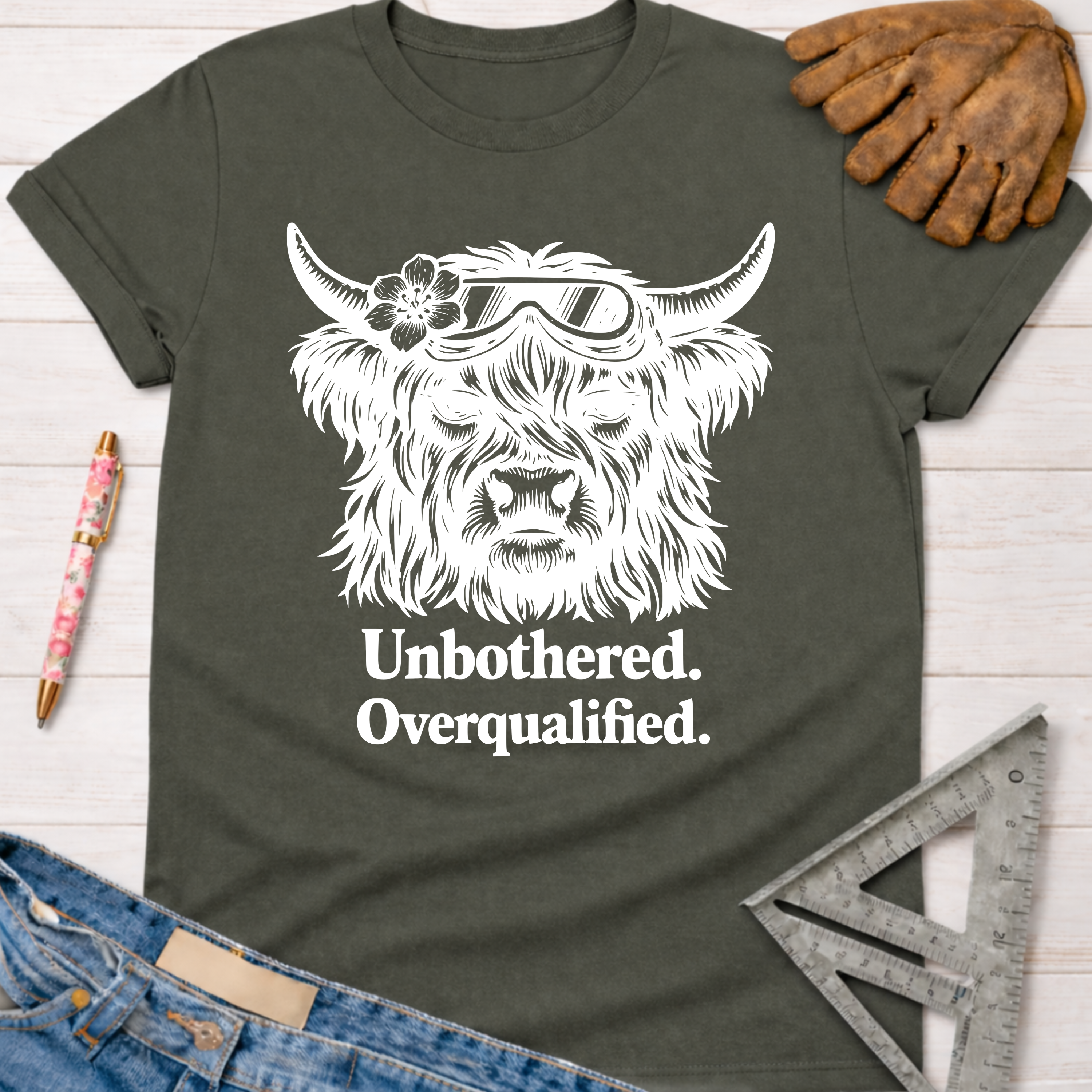 Women's funny woodworking graphic tee shirt for female woodworkers, makers, and DIY builders — Sawdust & Ponytails. Dark gray t-shirt with white "Unbothered. Overqualified." text and a Highland cow wearing goggles and a flower.