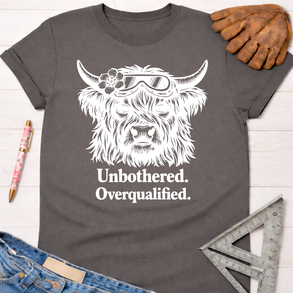 Women's funny woodworking graphic tee shirt for female woodworkers, makers, and DIY builders — Sawdust & Ponytails. Dark gray cotton tee with white graphic of a Highland cow wearing safety goggles and a flower, text "Unbothered. Overqualified.