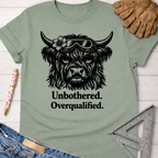 Women's funny woodworking graphic tee shirt for female woodworkers, makers, and DIY builders — Sawdust & Ponytails. Sage green cotton tee with "Unbothered. Overqualified." text and a Highland cow wearing goggles.