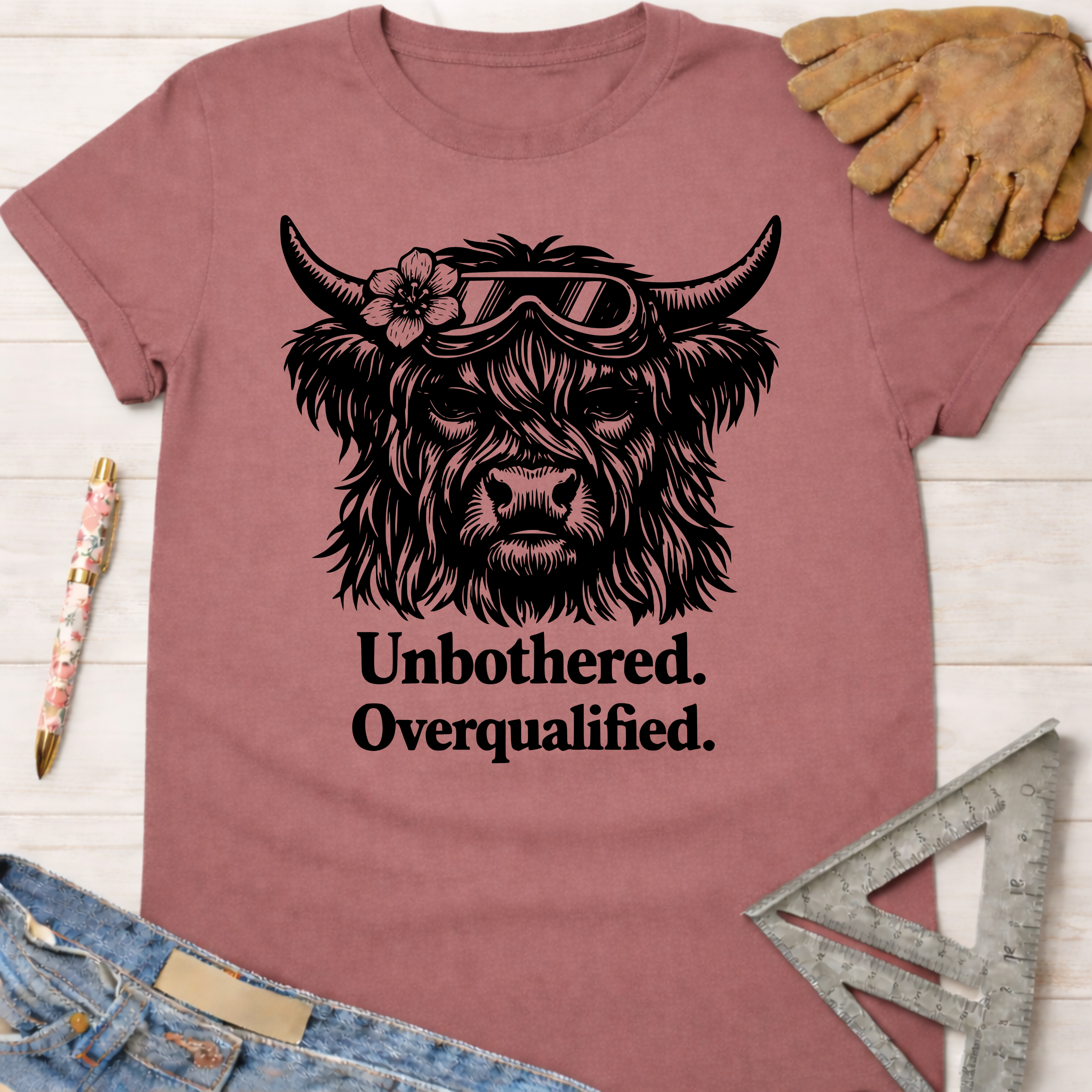 Women's funny woodworking graphic tee shirt for female woodworkers, makers, and DIY builders — Sawdust & Ponytails. Mauve t-shirt with a black graphic of a Highland cow wearing goggles and a flower, text reads "Unbothered. Overqualified.