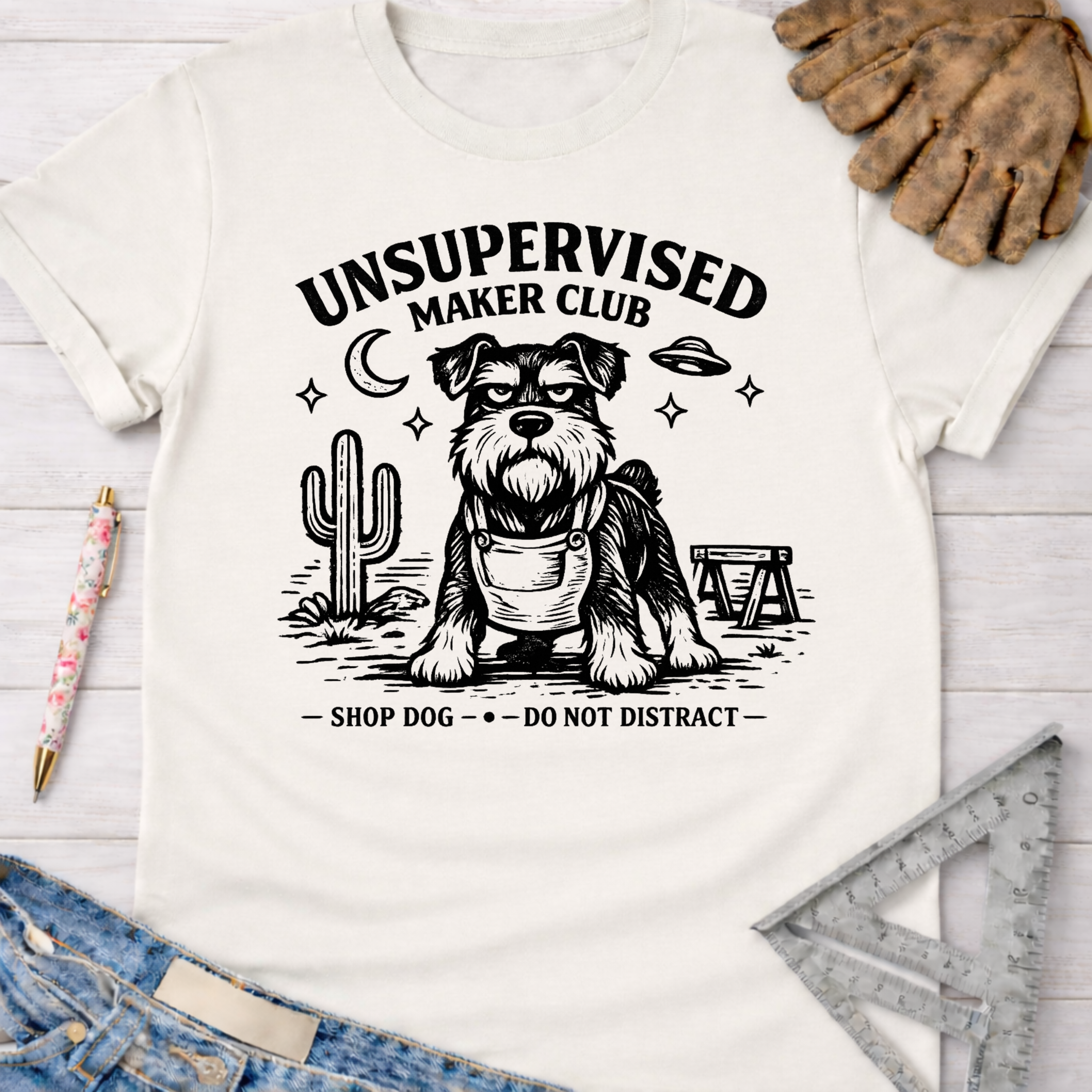 Women's funny woodworking graphic tee shirt for female woodworkers, makers, and DIY builders — Sawdust & Ponytails. White cotton tee with a black graphic of a schnauzer in overalls, "Unsupervised Maker Club" text, cactus, and UFO.