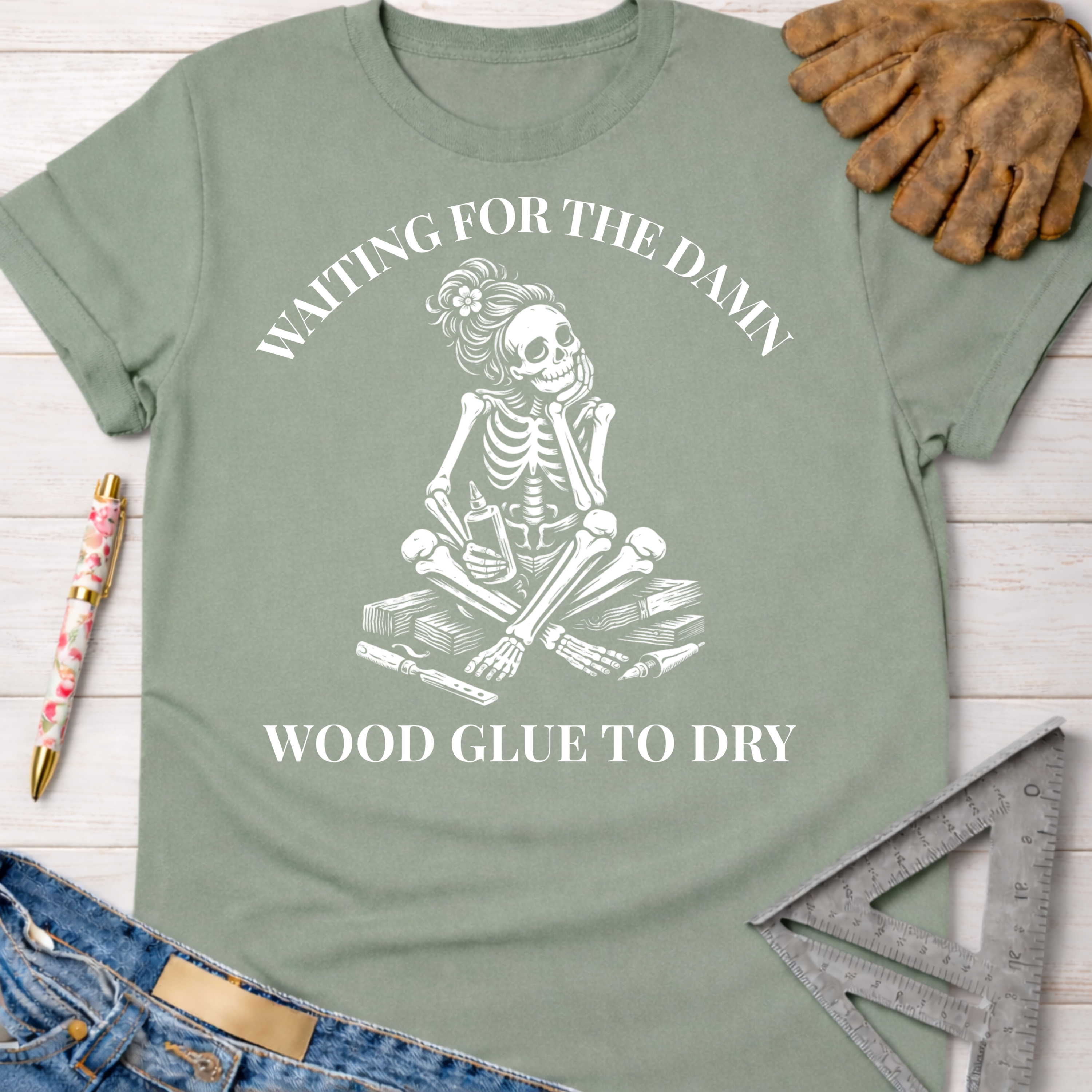 Women's funny woodworking graphic tee shirt for female woodworkers, makers, and DIY builders — Sawdust & Ponytails. Sage green t-shirt with a skeleton waiting for wood glue to dry.