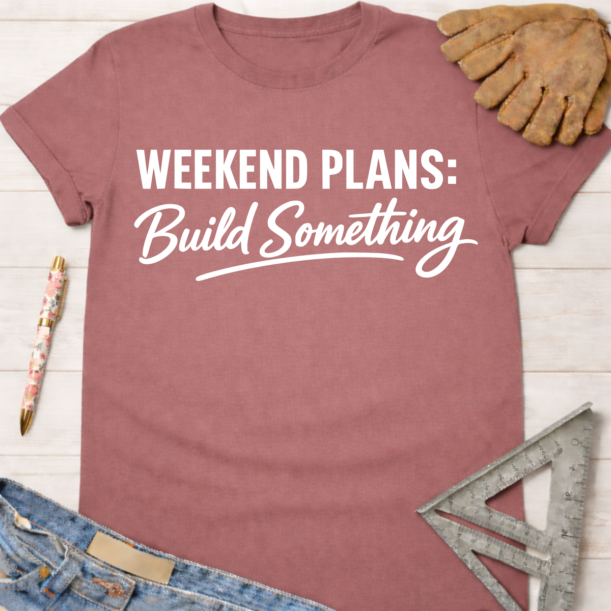 Women's funny woodworking graphic tee shirt for female woodworkers, makers, and DIY builders — Sawdust & Ponytails. Mauve cotton t-shirt with "Weekend Plans: Build Something" in white text, styled with jeans, a pen, work glove, and speed square.