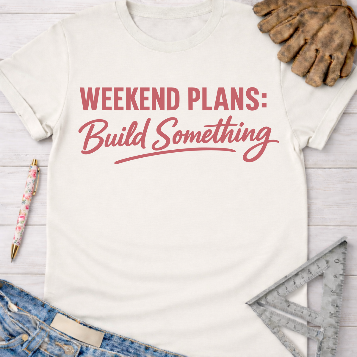 Women's funny woodworking graphic tee shirt for female woodworkers, makers, and DIY builders — Sawdust & Ponytails. Cream t-shirt with "Weekend Plans: Build Something" in red script, beside work gloves, a pen, jeans, and a ruler.