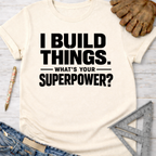 Women's funny woodworking graphic tee shirt for female woodworkers, makers, and DIY builders — Sawdust & Ponytails. Cream t-shirt with "I BUILD THINGS. WHAT'S YOUR SUPERPOWER?" in black text, styled with jeans, work gloves, and a ruler.
