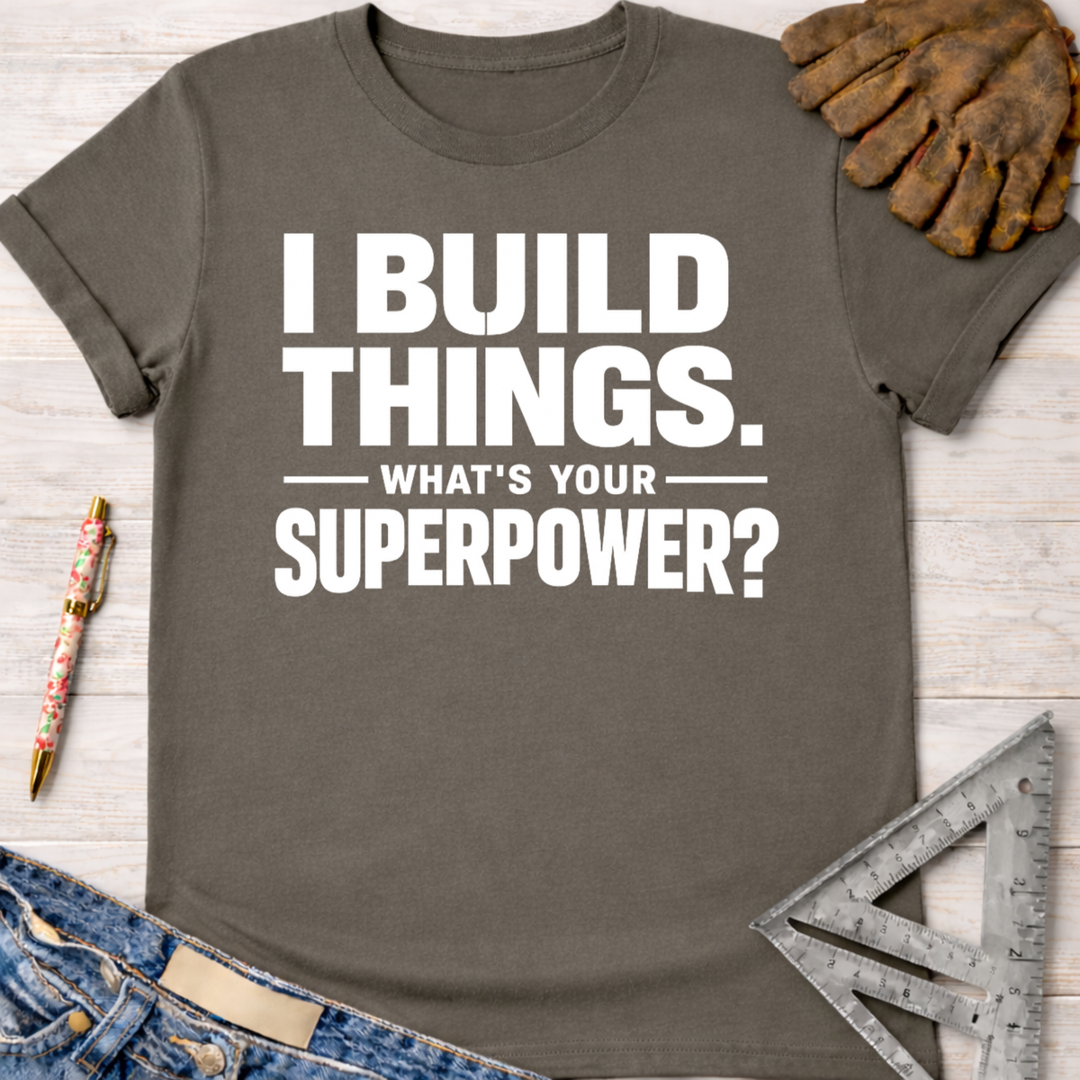 Charcoal gray women's "I Build Things. What's Your Superpower?" graphic tee, with work gloves and jeans.