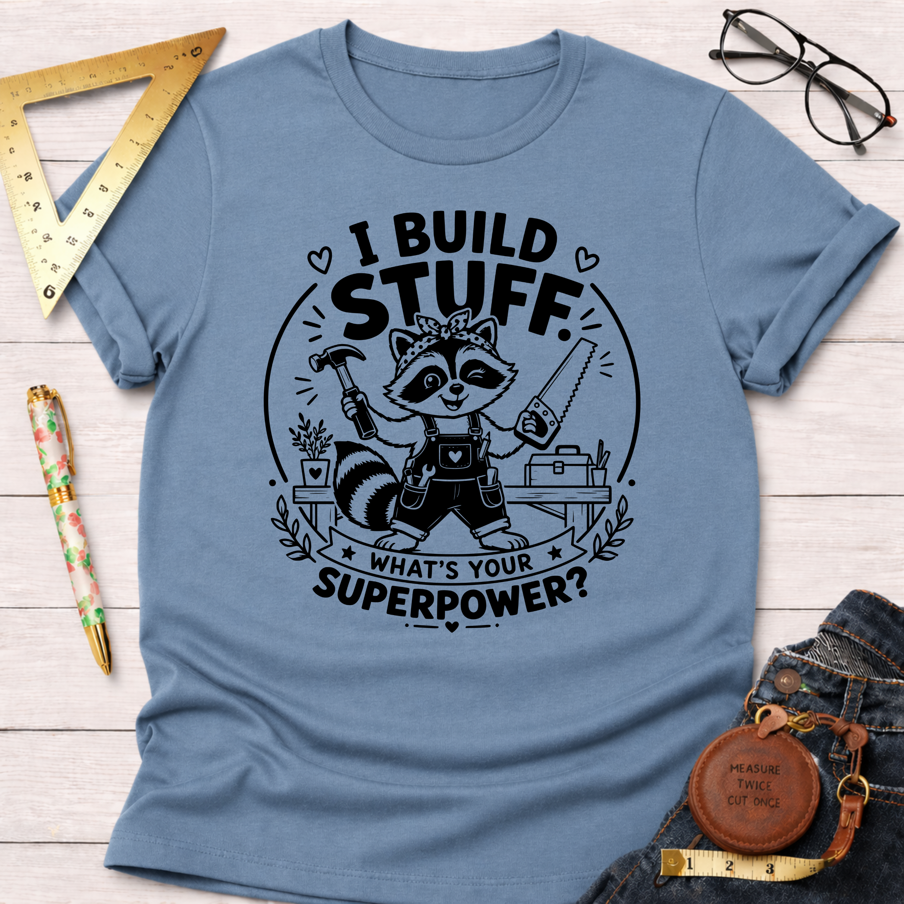 Light blue cotton t-shirt with a raccoon builder graphic holding a hammer and saw, "I BUILD STUFF. WHAT'S YOUR SUPERPOWER?" text.