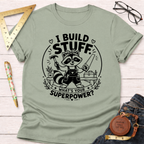 Sage green cotton t-shirt with a raccoon builder graphic holding a hammer and saw, "I Build Stuff. What's Your Superpower?" text.