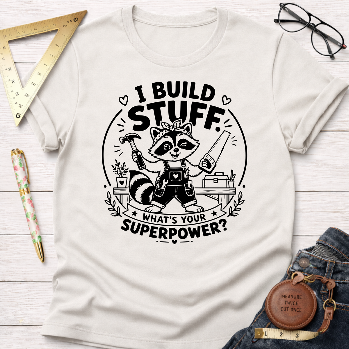 Cream cotton t-shirt with a black graphic of a raccoon builder holding a hammer and saw, text reads "I BUILD STUFF. WHAT'S YOUR SUPERPOWER?".