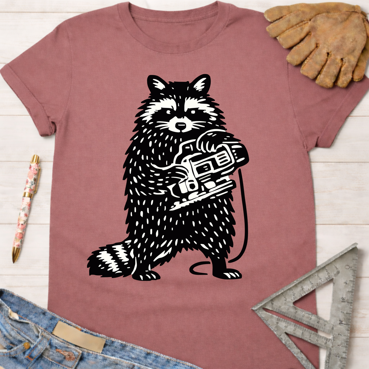 Women's funny woodworking graphic tee shirt for female woodworkers, makers, and DIY builders — Sawdust & Ponytails. Mauve t-shirt with a black and white raccoon holding a jigsaw.