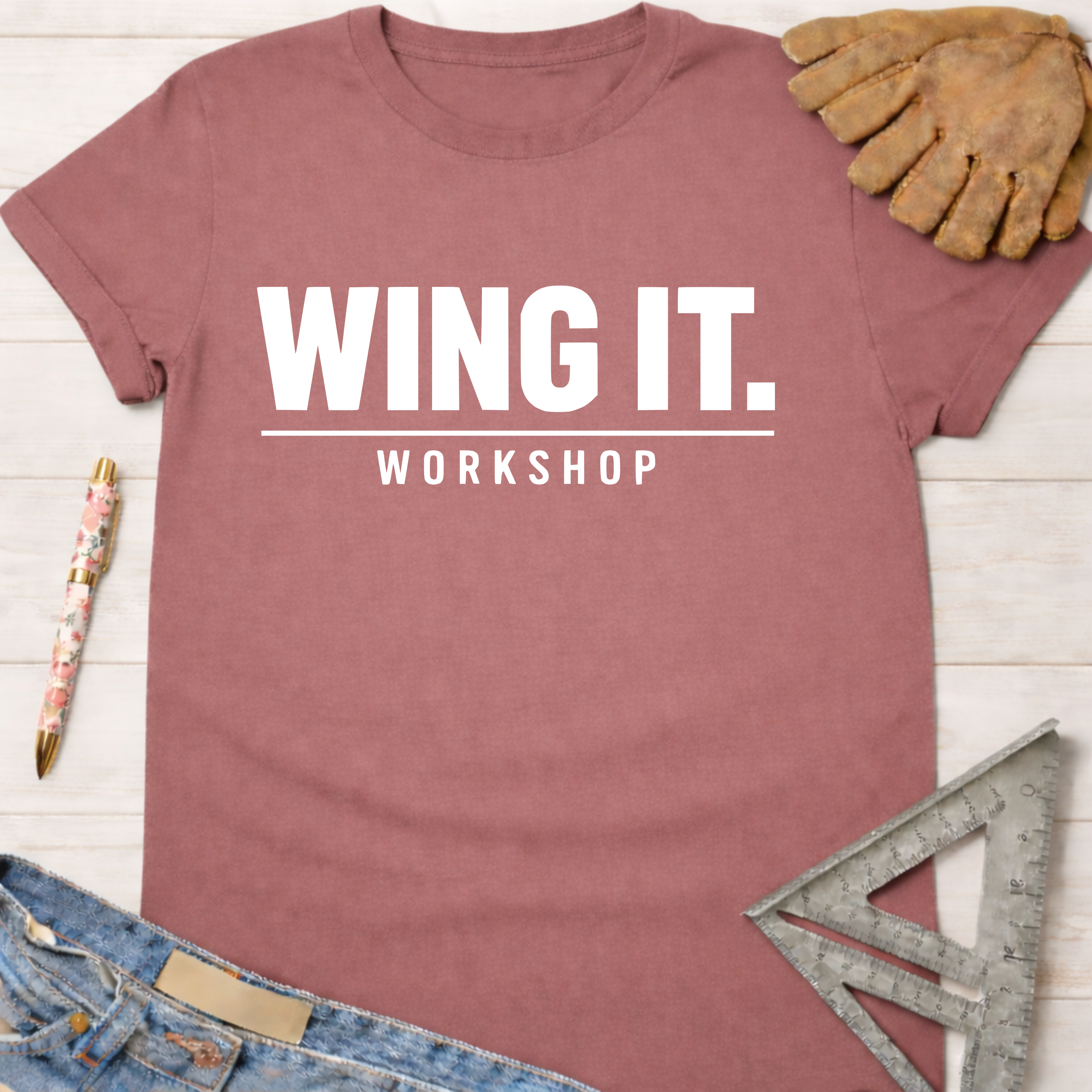 Women's funny woodworking graphic tee shirt for female woodworkers, makers, and DIY builders — Sawdust & Ponytails. Mauve t-shirt with "WING IT. WORKSHOP" in white text, next to work glove, pen, jeans, and speed square.